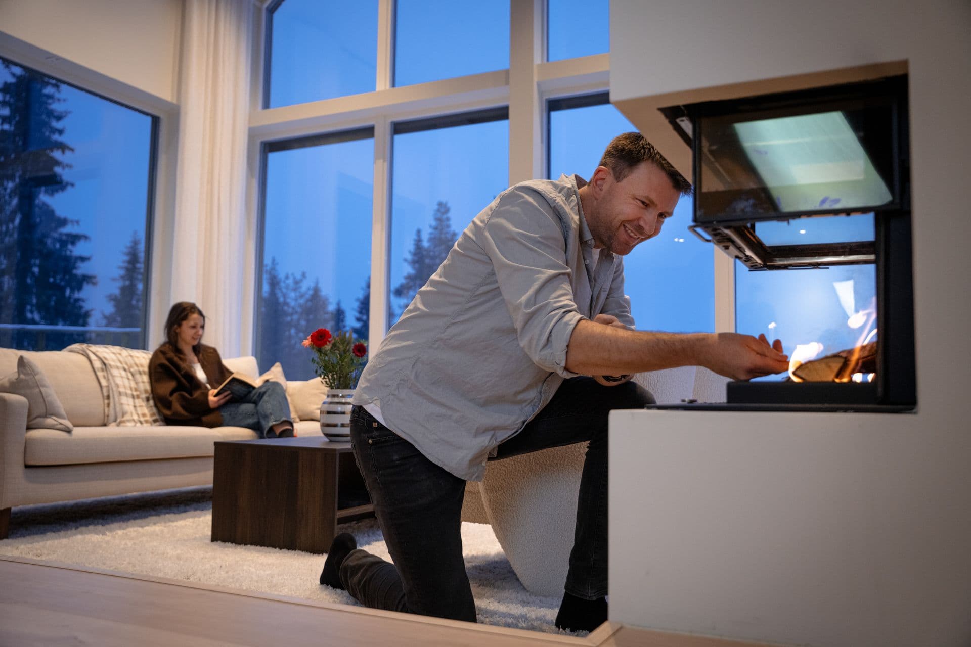 Mann som fyrer i peisen mens en kvinne sitter og leser i sofaen bak.