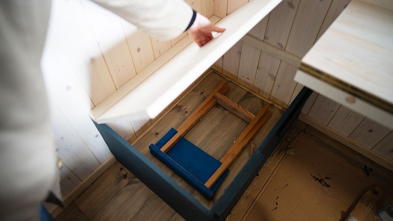 Hånd som holder lokket på en oppbevaringsboks åpen. I boken ligger det en krakk som er slått sammen.