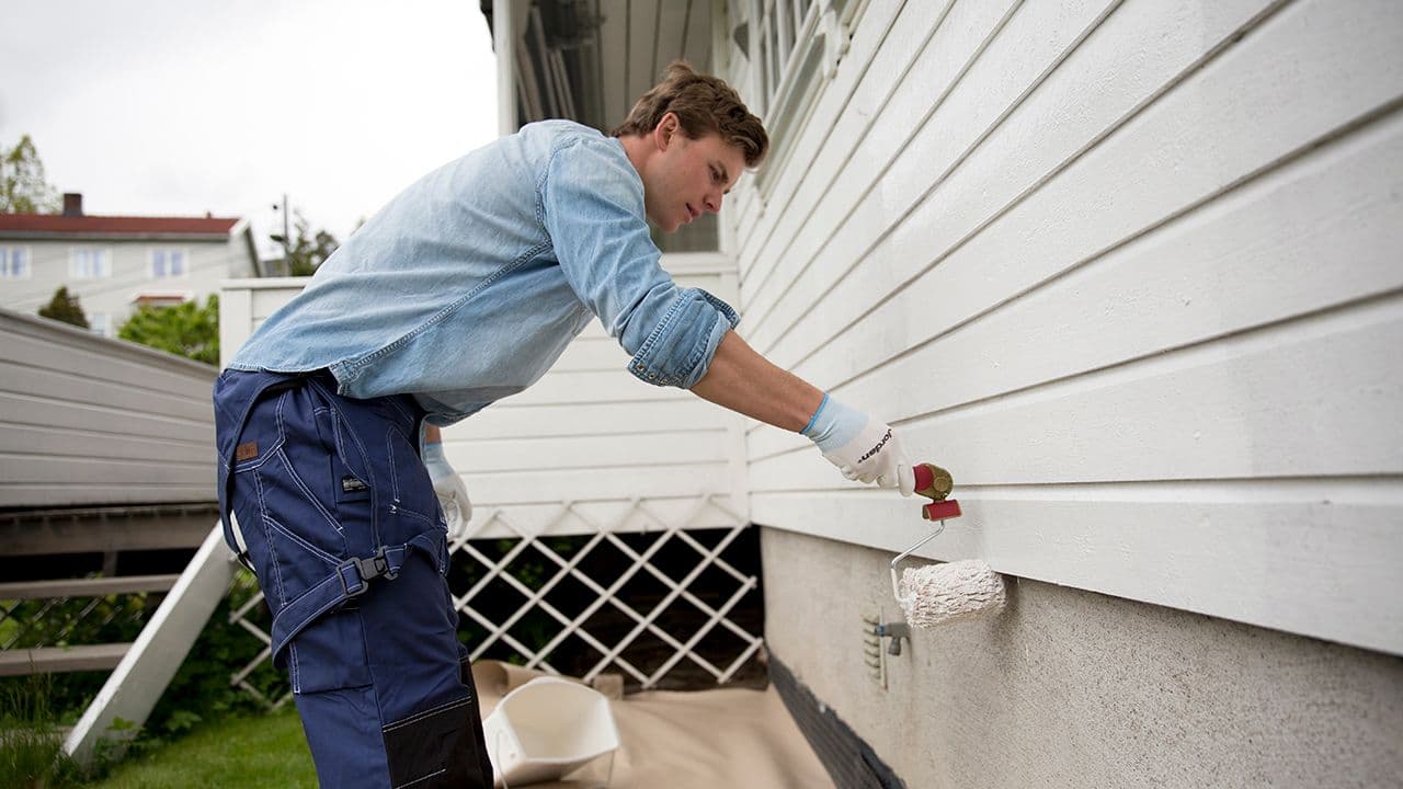 Mann som bruker rulle for å male et hus med hvit maling.