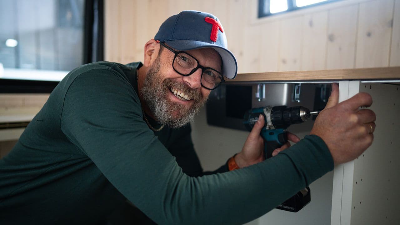 Mann som smiler mot kamera mens han bygger sammen kjøkkeninnredning i minihytte.
