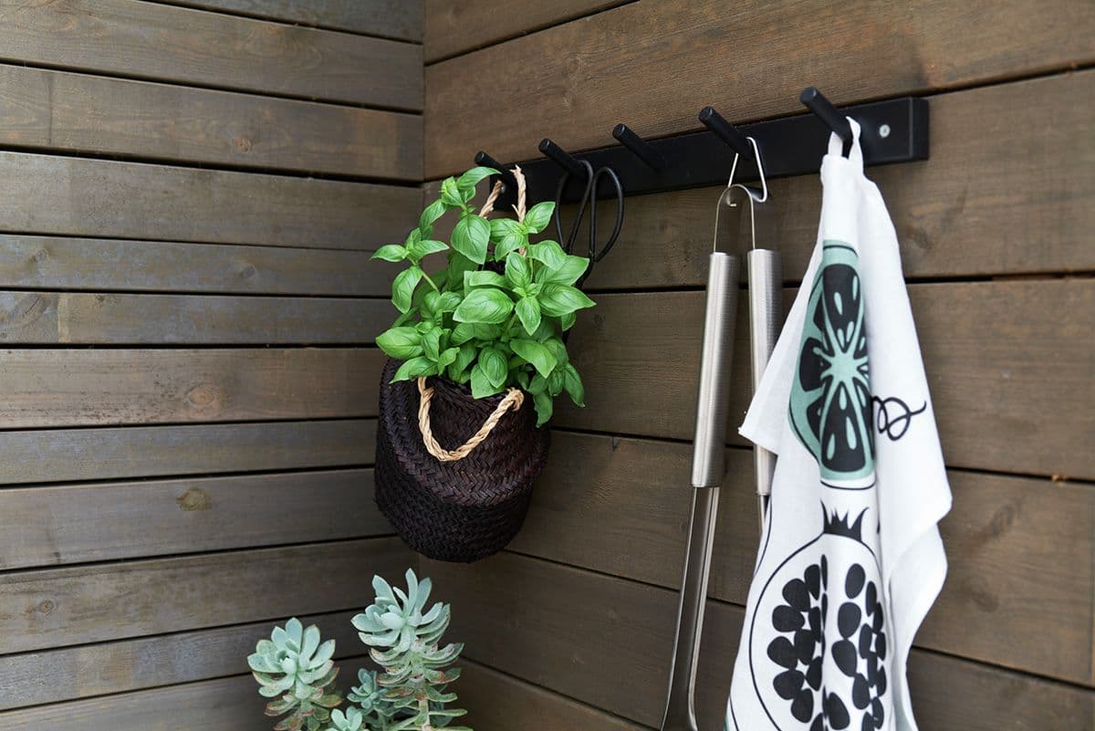 Husfasade med gråbeiset MøreRoyal-kledning i to ulike formater. På veggen henger en knaggrekke med grilltilbehør og urteplante.