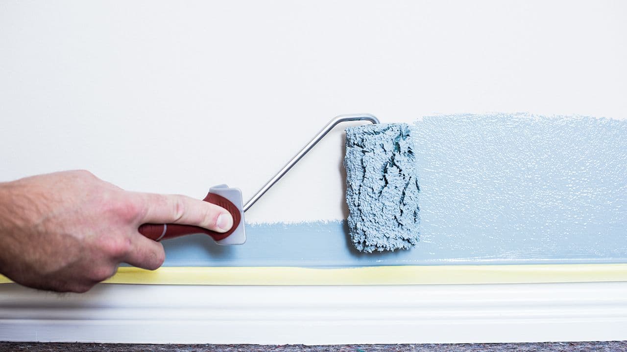 Maskeringsteip over gulvlist hvor en mann maler vegg inne med malingsrulle.