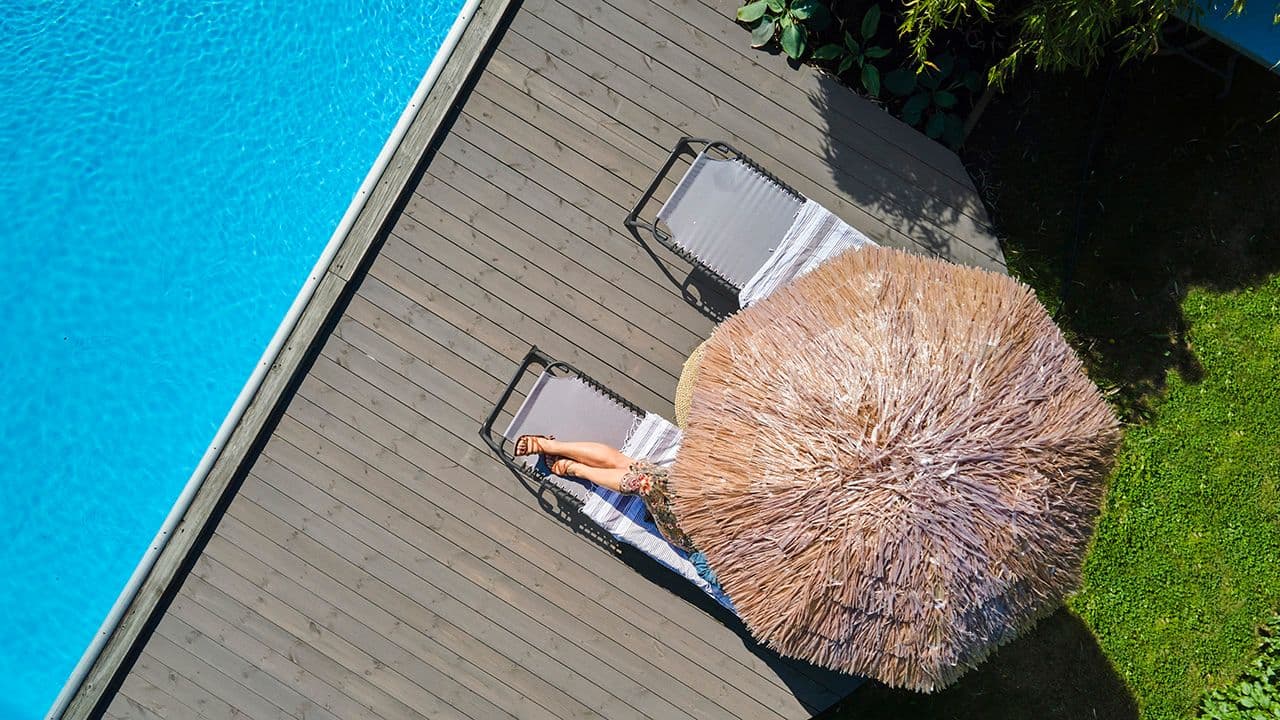 Person under parasoll på terrasse med vedlikeholdsfrie terrassebord ved basseng.