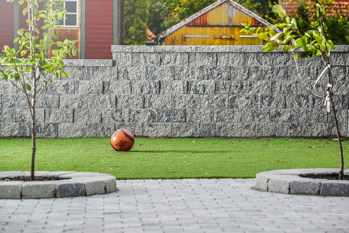 Mulitblokk støttemur med struktur i overflaten i hage med gress og belegningsstein.