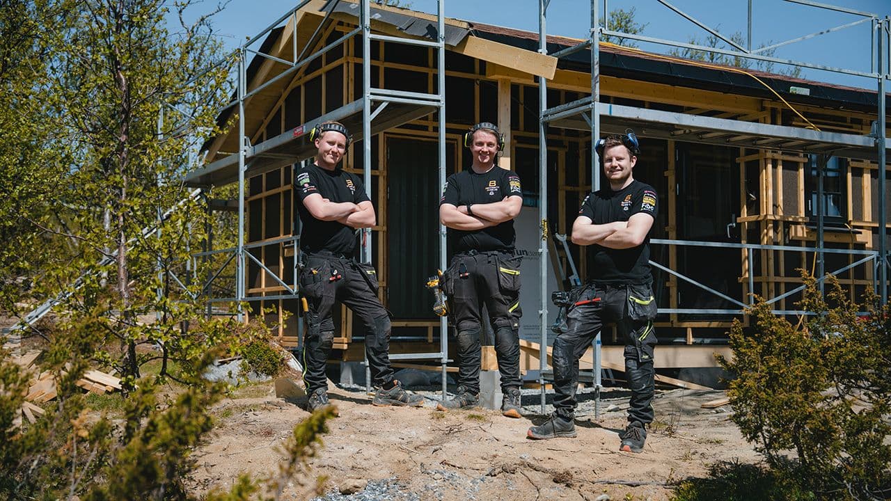 Tre håndverkere med arbeidsklær og hørselsvern foran et hus under renovering.