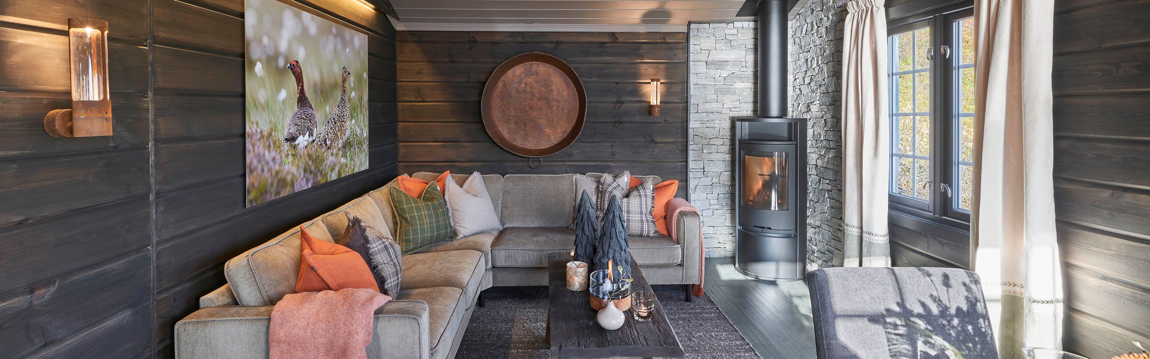 Hytteinteriør med liggende trepanel i stue som er beiset mørk og har grå sofa og peis i hjørnet.